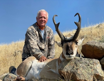 A seasoned hunter in camouflage with a grey hairstyle, proudly kneeling beside his antelope trophy against a hilltop backdrop.